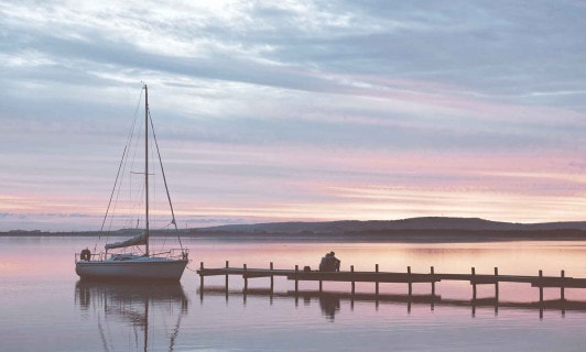Ein Boot auf einem See und zwei Menschen auf einem Steg im Sonnenuntergang.