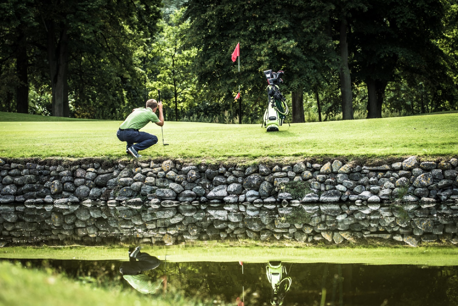 Ein Mann kniet auf einem Golfplatz am Rand des Ufers vor einem Teich.