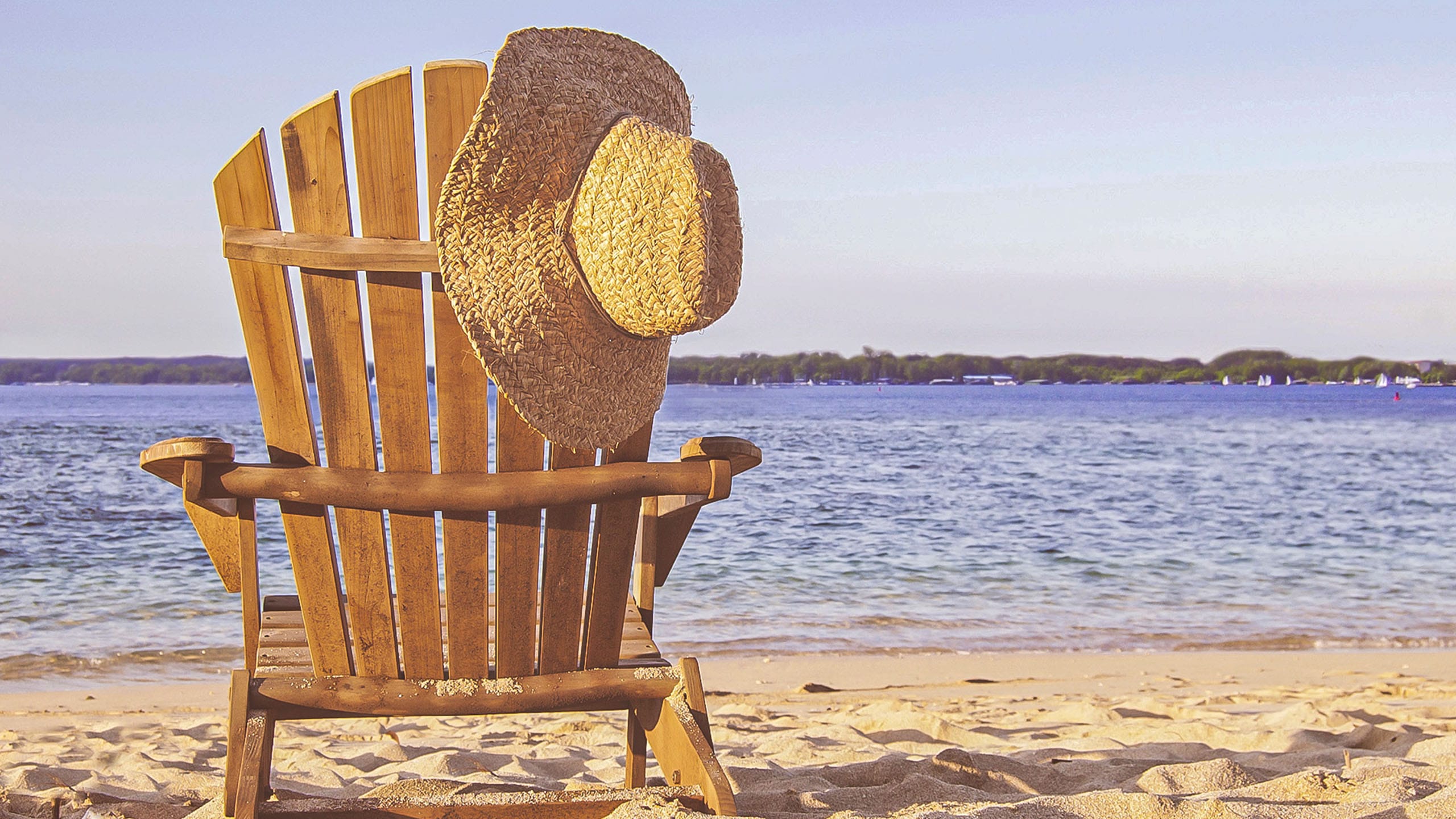 Ein Strandstuhl aus Holz am See Strand, auf dem ein Strohhut hängt.