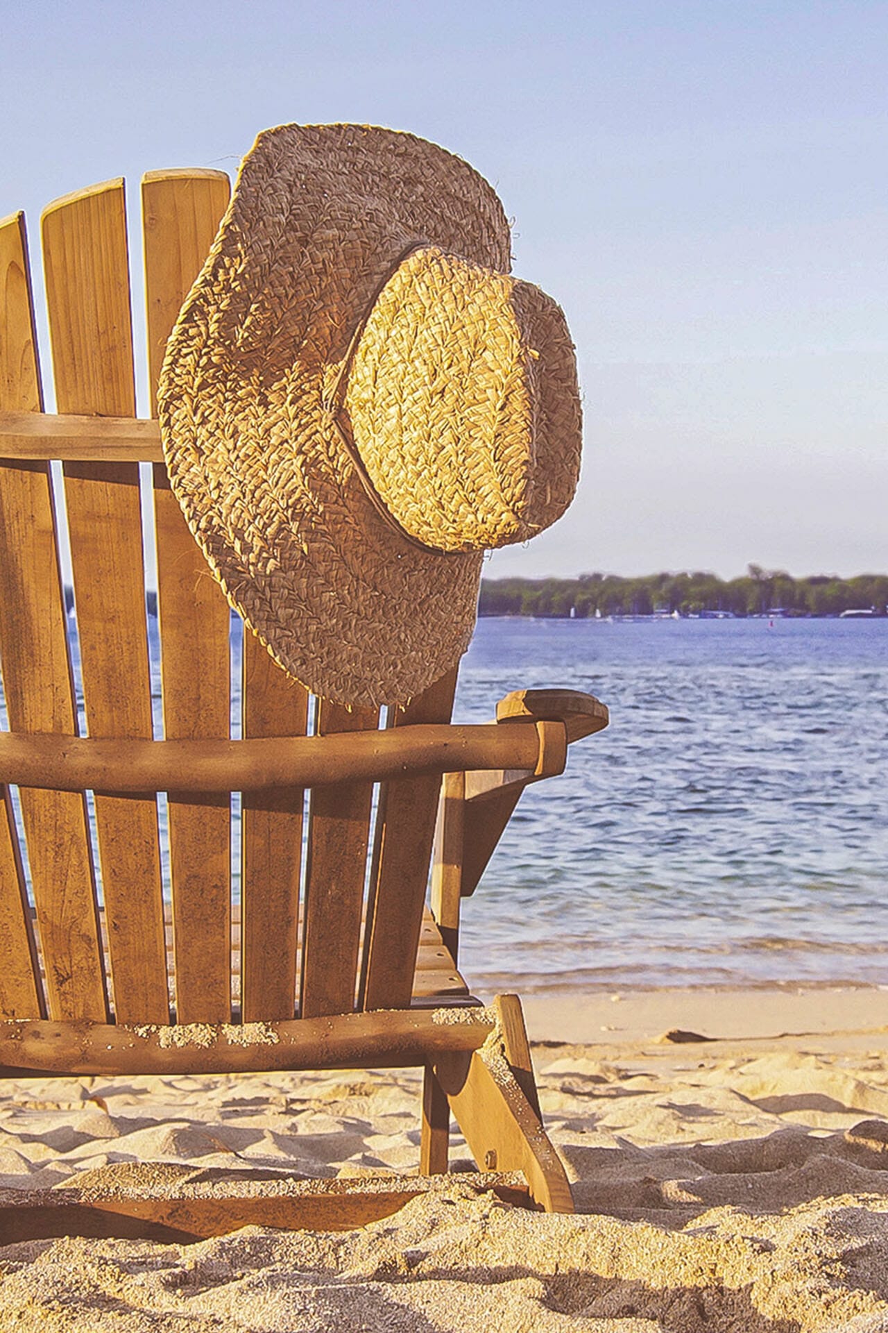 Ein Strandstuhl aus Holz am See Strand, auf dem ein Strohhut hängt.