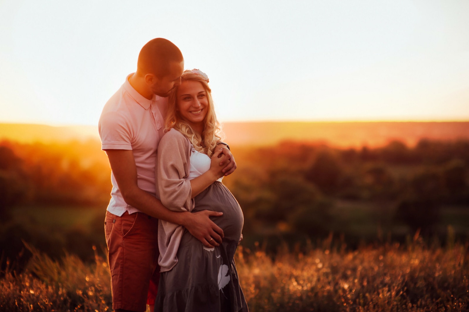 Eine schwangere Frau und ihr Mann stehen auf einer Wiese im Sonnenuntergang.